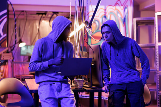 Twp hackers breaching cybersecurity system to gain illegal access. Man in hood showing computer virus code on laptop to malicious software developer and asking for advice