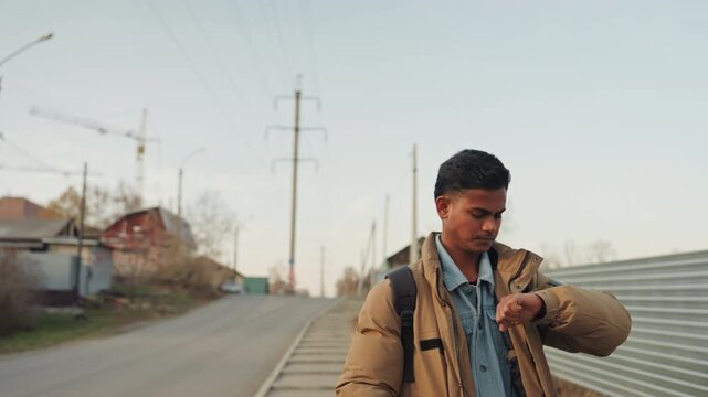 Young man on suburban street checking watch and backpack, quiet autumn light, cranes and metal fence in background, distant car passes, contemplative mood implying upcoming repair appointment