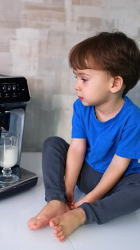 Beautiful Caucasian baby in blue t-shirt sits on the table near the coffee machine. Kid watches milk pouring into the cup from device.