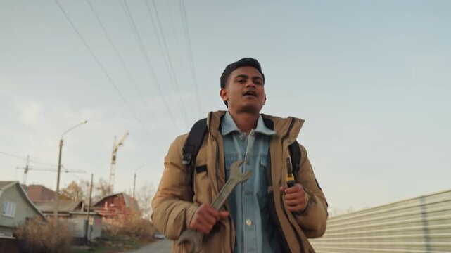 South asian man holding screwdriver and wrench on suburban street, backpack on shoulders, inspecting tool with focused expression, utility poles and houses in background, golden sunlight, candid