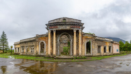 Fototapeta premium Abandoned Soviet era railway station in the mining town of Tkuarchal. Ruined neoclassical building with tall columns and arched windows under a cloudy sky in spring. Abkhazia.