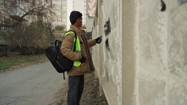 Worker inspecting vandalized concrete wall, backpack and reflective vest, gloved hands checking paint damage and graffiti, preparing tools to repair and repaint surface, overcast alley, community