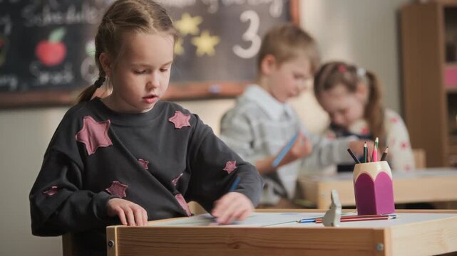 Medium shot of inspired Caucasian girl drawing with pencil while sitting at table in front of blackboard among children in kindergarten classroom
