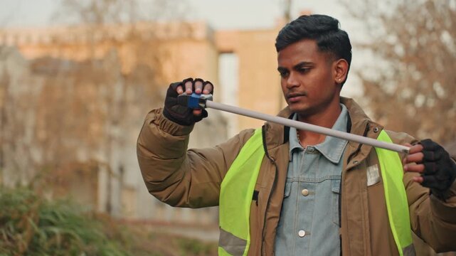 Worker measures plank with tape for fix and cut planning, concentrated expression near alley, highvisibility vest, gloved hands extend tape, careful length check under warm light