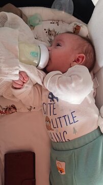 The newborn lies comfortably in a soft playpen and drinks milk from a bottle in a cozy home environment during feeding.
