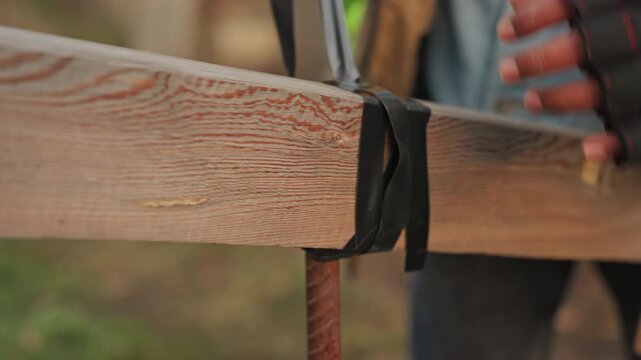 Closeup wood beam makeshift repair with tape, gloved hands wrapping black tape around cracked corner to stabilize plank, outdoor construction scene, improvised contractor action and emergency patching