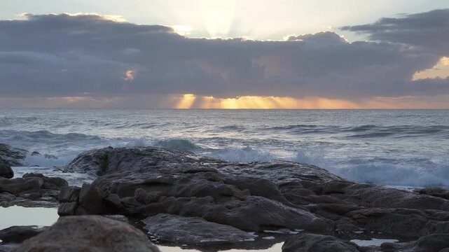 Dramatic sunrise as sunbeams pierce through foreboding clouds over a calm sea, illuminating waves as they crash against a rugged, rocky coastline at Ramsgate, South Coast.