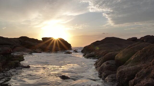 The sun rises through scattered clouds, over a rocky shore, casting golden rays over the rocks onto the water's surface at Ramsgate, South Coast.