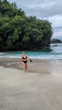 Happy Woman in Bikini Posing on Tropical Beach Holding Snorkeling Mask with Turquoise Ocean and Green Cliff in Background