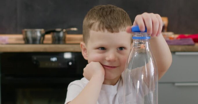 Young boy twists blue cap from clear bottle studying recycling step at table. Preschooler child lifts loose lid with fingers starting sorting task carefully slow motion