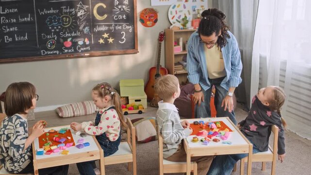 Handheld full shot of young biracial male kindergarten teacher joining Caucasian children shaping figures using colorful modeling clay and sitting at tables in pairs in cozy classroom with blackboard