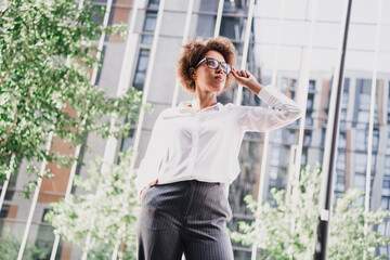 Fototapeta premium Confident mixed race woman in white shirt and grey trousers stands in front of modern city buildings for a stylish business lifestyle photo shoot