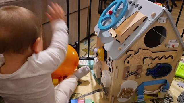 Little toddler playing with wooden busy board house educational toy developing fine motor skills and sensory exploration in a cozy home environment using Montessori method. 