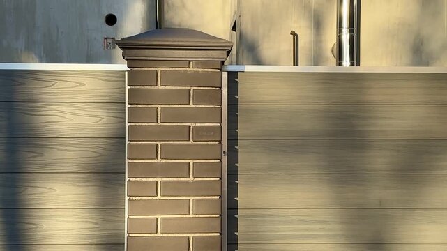 Sunlit brick column by cottage fence, warm golden hour light casts long tree shadows across weathered wooden panels, capstone.