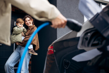 Family standing as father charges electric car at home, mother holding child. Embracing clean...