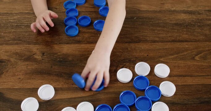 Boy sorts blue and white bottle caps by color on wooden table. Child slides plastic lids into groups building neat piles for recycling practice at home