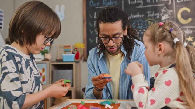 Medium slowmo shot of young biracial male teacher engaged in creative activity with Caucasian girl and boy sitting at table in kindergarten classroom and creating figures from modeling clay