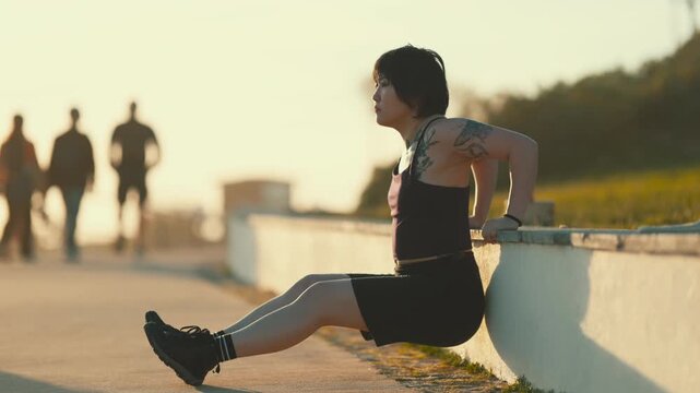 Tattooed asian woman performing triceps dips outdoor during sunset workout