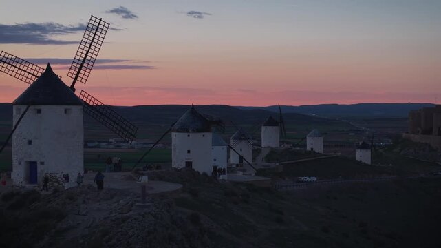 Iconic windmills of Consuegra in Castilla La Mancha, Spain, silhouetted against a beautiful sunset sky with vibrant pink and orange colors over the rolling hills