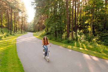 Fototapeta premium Happy woman rides bicycle along path in sunny forest. A young woman enjoys bike ride and relaxes in a park. Concepts of leisure, weekends, and transportation.