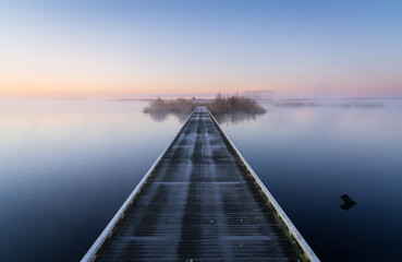 Fototapeta premium Wooden footbridge over a lake during a foggy and cold, spring morning in the Netherlands.