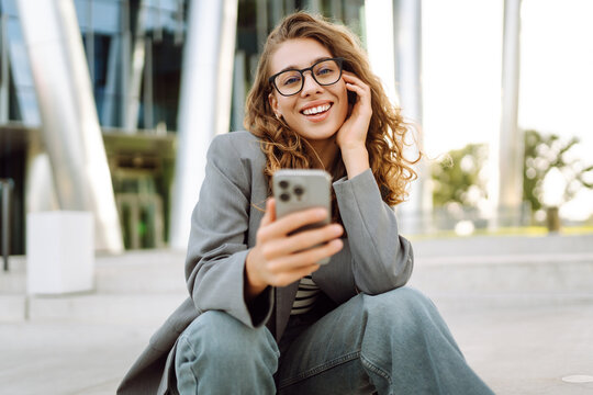 A young woman in stylish clothing, holding a phone, stands against the backdrop of a modern building. A businesswoman texts on her phone outdoors. Concepts: technology, modernity, business.