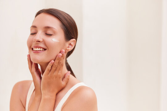 Natural beauty woman smiling joyfully with eyes closed and a cream swatch on her cheekbone, cradling her face with both hands during her skincare routine against a soft neutral background.