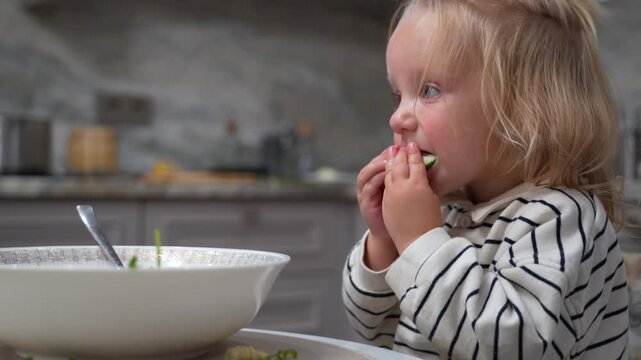 Young child experiences moment of hesitation and then tentatively begins to eat from bowl of food in kitchen