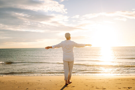 A gray-haired woman enjoys the seascape at sunset. A relaxed elderly woman strolls along a sandy beach, feeling free. Concepts for relaxation and retirement.