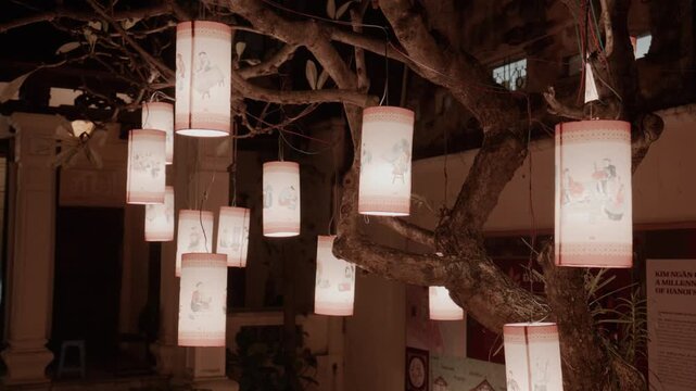 Traditional vietnamese paper lanterns with illustrations hanging from tree branches at night illuminating dark in ancient city temple in Hanoi, Vietnam