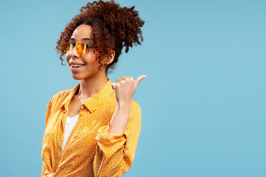 Smiling young black woman pointing sideways with thumb on blue background