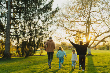Fototapeta premium Parents and their two children run joyfully across a grassy lawn under a large tree at sunset. Happy family recreation and active lifestyle concept.