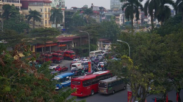 Hectic traffic jam with cars, buses, motorbikes moving through busy street in downtown Hanoi, Vietnam, surrounded by lush green trees and urban architecture