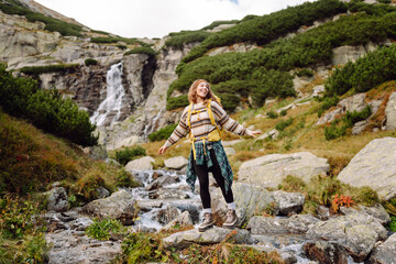 A woman enjoys her time exploring the mountains with a backpack. She stands near a waterfall and looks happy while surrounded by green plants and rocks. Hiking concept. © maxbelchenko