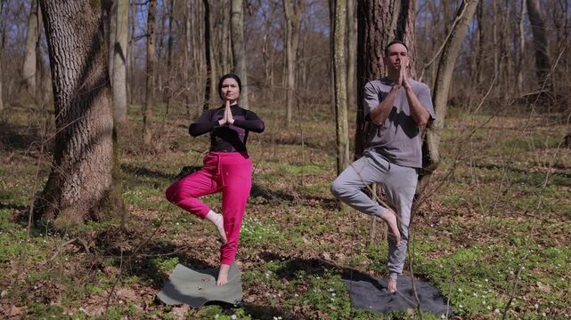 Peaceful couple doing yoga vrikshasana pose together in the middle of a sunny spring forest. Man and woman meditating, standing on one leg for balance, harmony, and wellness