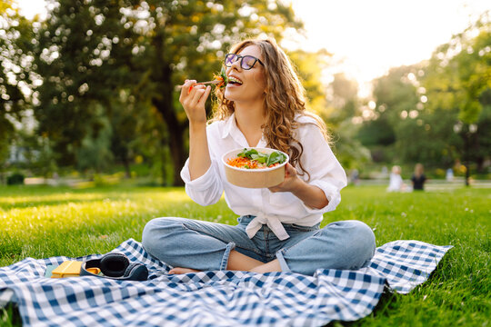 A cheerful woman wearing glasses sits on a mat in a park, eating a salad on a sunny day. A young woman enjoys lunch outdoors. Concept: relaxation, snacking, outdoors.