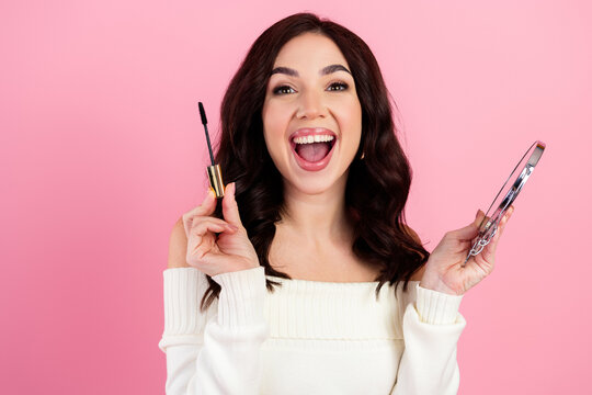 Smiling woman applying mascara while holding a compact mirror, joyful beauty routine and makeup tutorial moment on pink background