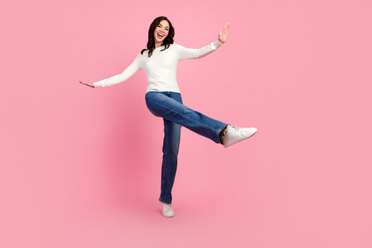 Happy young woman in white sweater and jeans balancing playfully on one leg against a pink studio background, smiling and radiating carefree, energetic lifestyle and casual fashion
