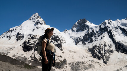 A woman hiker with a backpack stands in the snowy mountains during an acclimatization trek. The side view portrait of a female backpacker with hiking gear, looking up at the sunlight. © Соня Монштейн