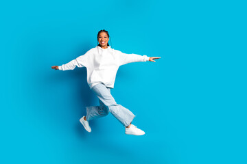 Fototapeta premium Young woman in white hoodie leaps with joy against blue background in a casual fashion photoshoot