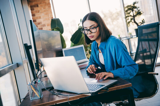 Female programmer works at a modern loft office using a laptop and monitor during daylight in a focused and productive moment