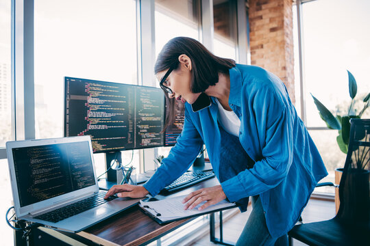 Busy programmer woman at a modern loft office debugging code on multiple screens while collaborators brainstorm new software project