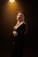 Fototapeta premium Pregnant woman in elegant black dress, artistic studio portrait with golden backlight