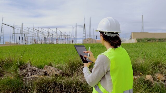 Female power engineer inspecting electrical substation infrastructure using a digital tablet, monitoring energy grids and network systems for efficiency and safety