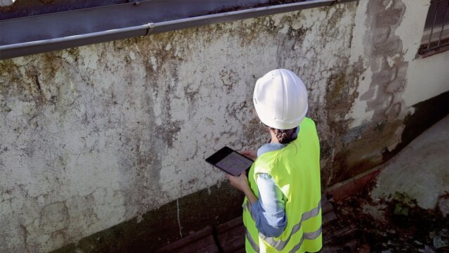 Engineer in hardhat and hi-vis vest documenting severe water damage, rising damp and mold on aged exterior wall using a tablet during on-site building inspection and assessment