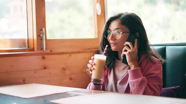 4K shot of Indian teenager girl talking on call and drink cold coffee on table at cafe at Manali, India. Social media addiction in youth. Technology, internet, communication and people concept.
