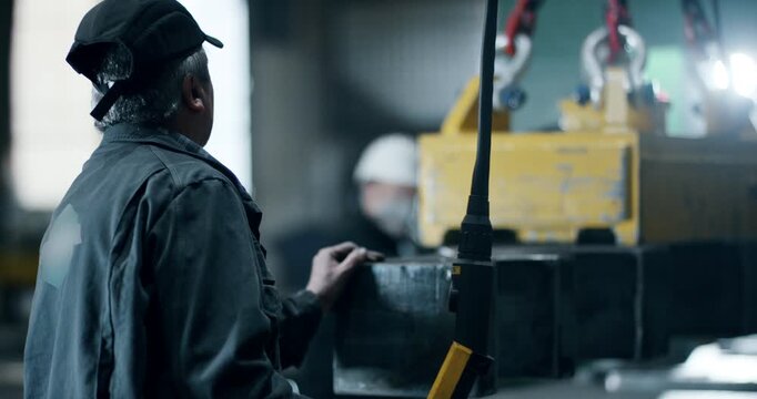 Rear view of a factory worker operating a magnetic lifter to move heavy steel parts in a workshop