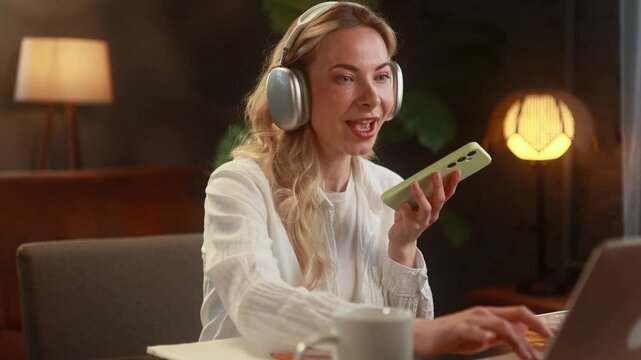 Young woman working on her laptop while dictating voice notes on her smartphone, speaking and organizing tasks using an audio planner during a focused remote work session at home