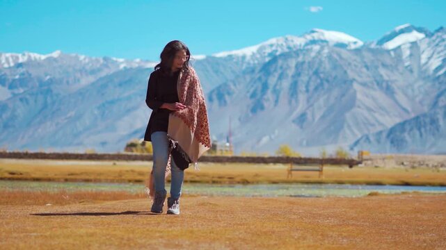 Young Indian woman wearing shawl walk on meadow against snow capped Himalaya mountains in Zanskar, Ladakh, India. Confident woman walking in beautiful mountains. Travel concept.