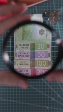 Vertical close-up of a stack of Ukrainian Hryvnia banknotes. A magnifying glass moves over the stack, highlighting the denomination text and numbers.